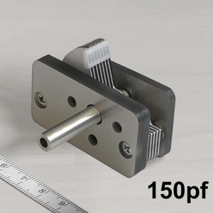 Puede incluir: Un componente electrónico plateado con una varilla cilíndrica de metal que se extiende desde una base rectangular. El componente tiene múltiples cables en blanco y negro y el texto "150pf" es visible.