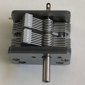 Puede incluir: Un condensador variable plateado y negro con un eje metálico y múltiples puntos de conexión. El dispositivo tiene una serie de placas metálicas paralelas y dos conectores blancos con múltiples pines. La parte superior tiene una pequeña pestaña metálica.