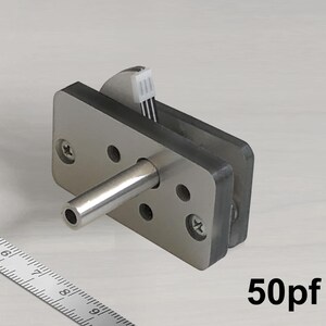 Puede incluir: Componente electrónico plateado con una varilla cilíndrica metálica y una base rectangular con múltiples orificios, asegurada con tornillos. El texto "50pf" está impreso en negro. Probablemente sea un condensador.