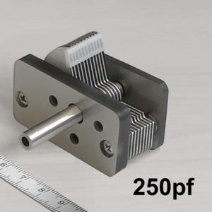 Puede incluir: Un condensador variable plateado con un rotor de plástico blanco y un eje de metal. El condensador tiene una capacidad de 250pF.