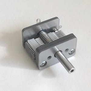 以下が含まれることがあります： 金属製のチューニングノブが付いた銀色の可変コンデンサー。コンデンサーは、透明なプラスチック製のハウジングを通して見える一連の金属プレートを持っています。