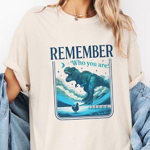 May include: Ivory-colored t-shirt with a graphic design featuring a teal dinosaur, a chicken, and the words "REMEMBER Who you are!" in navy blue. The design is framed within a rectangular border. The shirt is worn with a denim jacket.
