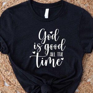 May include: Black short sleeve t-shirt featuring a white, cursive text graphic that reads, 'God is good all the time.'  Small heart shapes flank the text. The shirt is displayed on a woven surface.