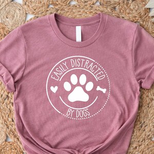 May include: A mauve t-shirt with a white circle graphic that says "Easily Distracted by Dogs" with a paw print and bone inside the circle.