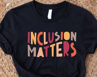 May include: Black t-shirt with the text "INCLUSION MATTERS" in colorful, retro-style lettering. The shirt is laid flat on a woven background.