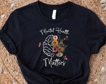 Puede incluir: Camiseta negra con un contorno blanco de un cerebro con flores en su interior y el texto "Mental Health Matters" debajo.