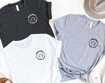 May include: Three t-shirts in charcoal, white, and light gray, each featuring a circular graphic with the text "INVISIBLE ILLNESS CLUB." The shirts are laid out with a pair of sunglasses, sandals, a hat, and sneakers.