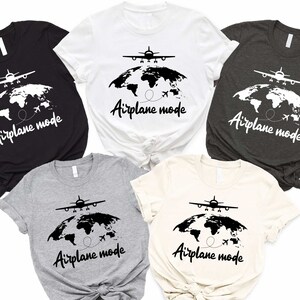 May include: Five short-sleeve t-shirts in various colors, including black, white, gray, and beige, feature a graphic design of a plane flying over a world map. The text 'Airplane mode' is printed below the graphic on each shirt. The shirts are arranged on a white background.  The design is simple and stylish, suitable for travel enthusiasts.