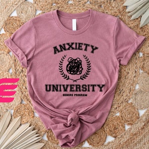 May include: A pink t-shirt with black text that reads "Anxiety University Honors Program". The text is in a circle with a wreath around it. There is a black doodle of a tangled mess in the center of the circle.