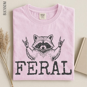 A pink t-shirt with a graphic design of a raccoon making a rock and roll sign, accompanied by the word "FERAL" printed below.