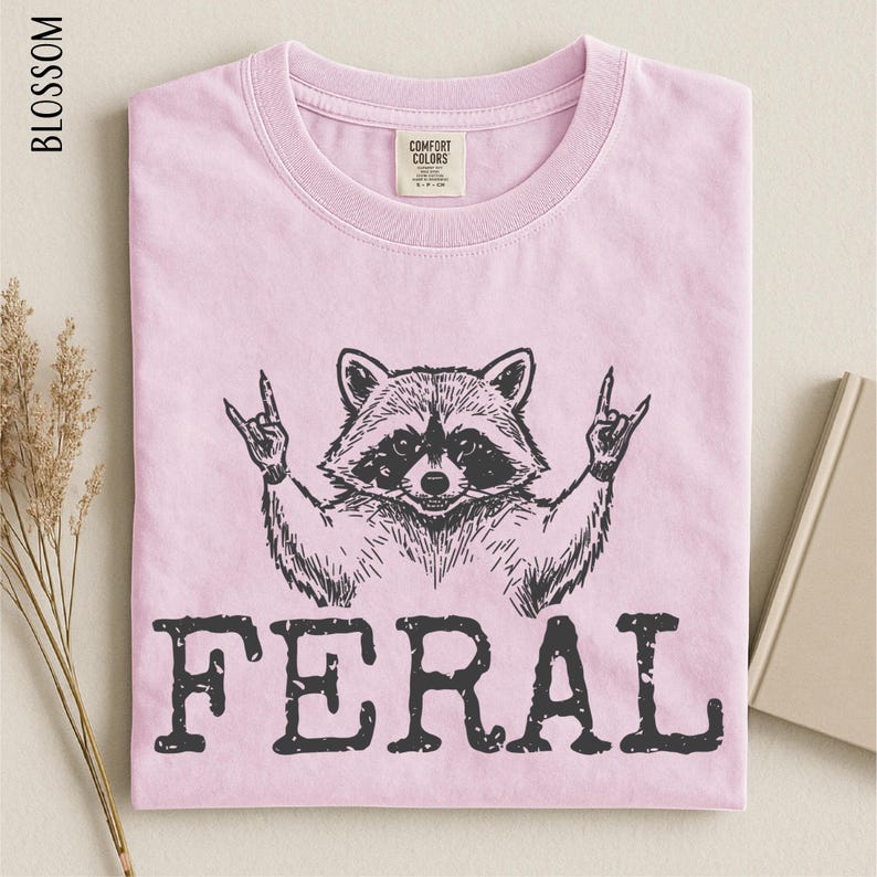 A pink t-shirt with a graphic design of a raccoon making a rock and roll sign, accompanied by the word "FERAL" printed below.
