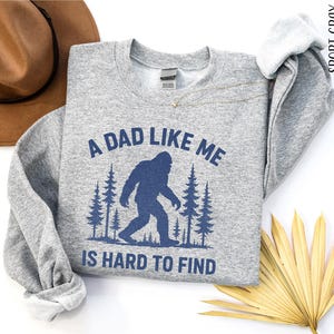 a gray sweatshirt with a graphic design of a Sasquatch silhouette and the text "A DAD LIKE ME IS HARD TO FIND" printed on it. The sweatshirt is laid out on a white background, accompanied by a brown hat, a fan, and some palm fronds.