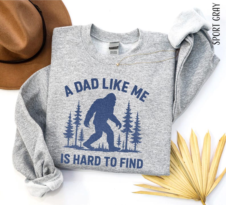a gray sweatshirt with a graphic design of a Sasquatch silhouette and the text "A DAD LIKE ME IS HARD TO FIND" printed on it. The sweatshirt is laid out on a white background, accompanied by a brown hat, a fan, and some palm fronds.