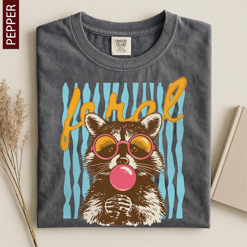 A gray t-shirt with a graphic design featuring a raccoon wearing pink bubblegum and sunglasses, along with the word "Raccoon" written above it.
