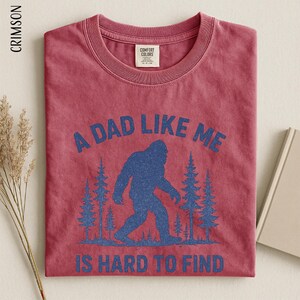 A red t-shirt with a graphic design depicting a blue Sasquatch silhouette and the text "A DAD LIKE ME IS HARD TO FIND" printed on it.