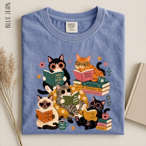 May include: A blue jean colored t-shirt featuring a graphic of five cats reading books. The cats wear glasses and are surrounded by stacks of books with titles like Feline Fiction, Cat Tales, MeoWoirs, and How to Relax, along with small coffee mugs.