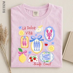 a pink t-shirt with a vibrant graphic design depicting various food items and icons, including fish, lemons, and a lobster, along with the text "La Dolce Vita" and "Analfi Coast".
