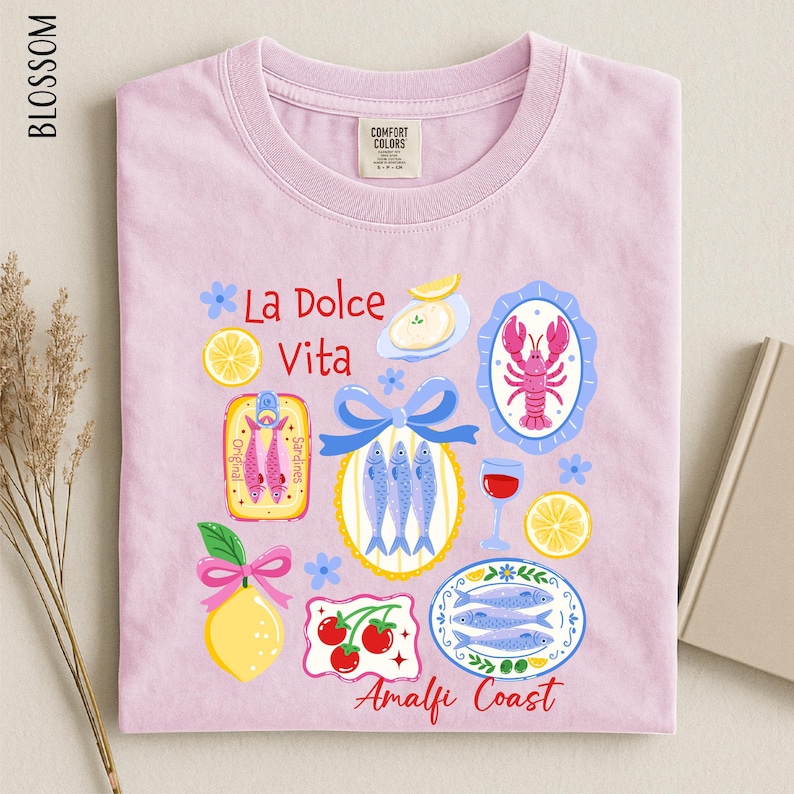 a pink t-shirt with a vibrant graphic design depicting various food items and icons, including fish, lemons, and a lobster, along with the text "La Dolce Vita" and "Analfi Coast".