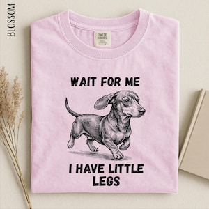 A pink t-shirt with a black and white illustration of a dachshund dog and the text "WAIT FOR ME I HAVE LITTLE LEGS" printed on it.