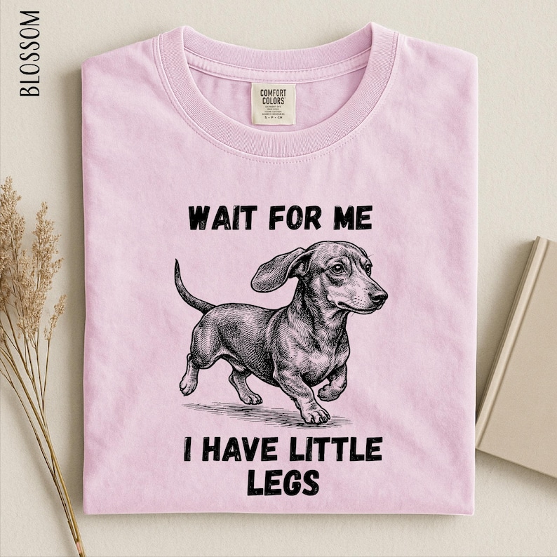 A pink t-shirt with a black and white illustration of a dachshund dog and the text "WAIT FOR ME I HAVE LITTLE LEGS" printed on it.