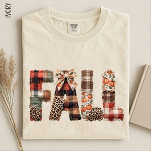 May include: An ivory t-shirt featuring the word FALL in large, block-style letters. Each letter is composed of various fabric patches including red and black plaid, leopard print, and pumpkin patterns. A decorative fabric bow sits atop the letter A.