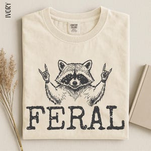 a t-shirt with a graphic design of a raccoon making a rock and roll gesture, accompanied by the word "FERAL" printed below.