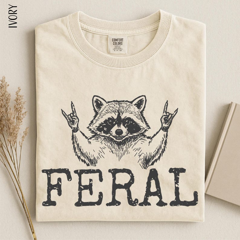 a t-shirt with a graphic design of a raccoon making a rock and roll gesture, accompanied by the word "FERAL" printed below.