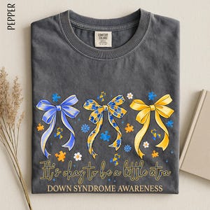 May include: A dark gray t-shirt with three decorative bows in blue, yellow, and gold, each adorned with puzzle pieces and flowers. The text "It's okay to be a little extra" and "Down Syndrome Awareness" are printed below the bows.