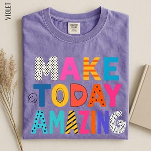 A purple t-shirt with the text "MAKE TODAY AMAZING" printed on it, along with various colorful and playful lettering designs.