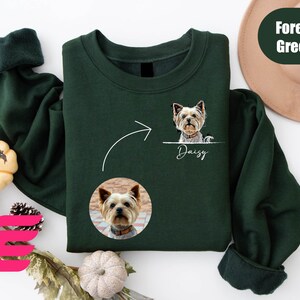K&ouml;nnte beinhalten: Ein dunkelgr&uuml;nes Sweatshirt mit einem wei&szlig;en Pfeil, der nach links zeigt, und einem wei&szlig;en Kreis mit einem Hundegesicht darin. Der Text "Daisy" befindet sich unter dem Kreis. Das Sweatshirt hat einen gerippten &Auml;rmel und einen Rundhalsausschnitt.
