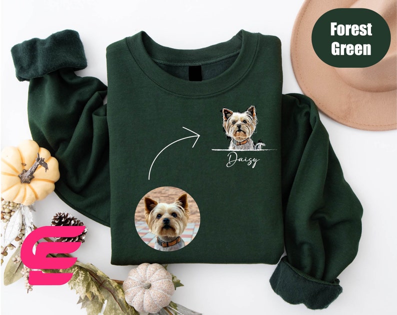 K&ouml;nnte beinhalten: Ein dunkelgr&uuml;nes Sweatshirt mit einem wei&szlig;en Pfeil, der nach links zeigt, und einem wei&szlig;en Kreis mit einem Hundegesicht darin. Der Text "Daisy" befindet sich unter dem Kreis. Das Sweatshirt hat einen gerippten &Auml;rmel und einen Rundhalsausschnitt.