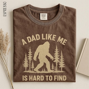 A brown t-shirt with a graphic design depicting a Sasquatch silhouette and the text "A DAD LIKE ME IS HARD TO FIND" printed on it.