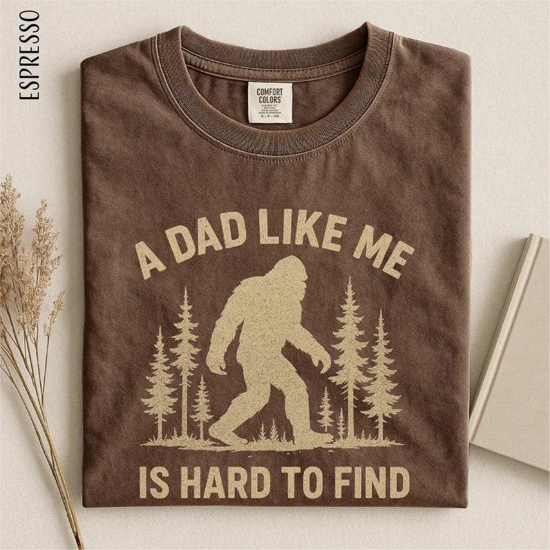 A brown t-shirt with a graphic design depicting a Sasquatch silhouette and the text "A DAD LIKE ME IS HARD TO FIND" printed on it.