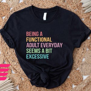 May include: Black t-shirt with the text "Being a Functional Adult Everyday Seems a Bit Excessive" in rainbow colors.