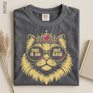 May include: Dark gray t-shirt with a gold cat graphic wearing a crown and sunglasses. The sunglasses have the words "THIS IS AN OUT RAGE" printed on them. The shirt is made of a soft material and has a Comfort Colors tag.