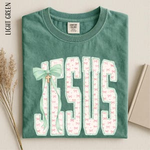 A green t-shirt with the word "JESUS" printed on it, along with a bow and a cross. The t-shirt is folded and placed on a surface, with some dried flowers and a book visible in the background.