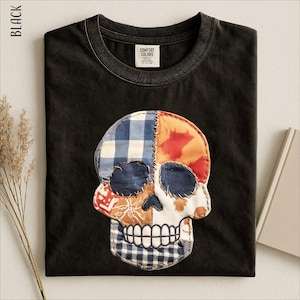May include: A black crew neck t-shirt featuring a large, central skull graphic made of various fabric patches. The skull design includes blue and white gingham, solid navy, and orange patterned sections, all joined by visible decorative stitching.