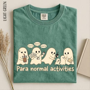 May include: A light green crew neck t-shirt featuring a graphic of five cartoon ghosts. The ghosts are holding items like a drink, documents, a stopwatch, and a walkie-talkie. The text "Para normal activities" and "d-a-t-a" is printed below.