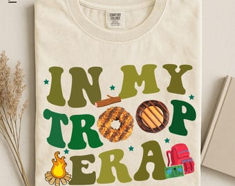 May include: An ivory t-shirt featuring the text "IN MY TROOP ERA" in large green letters. The graphic includes a small brown log, a coconut-topped cookie, a chocolate-covered cookie, a campfire, a red backpack, and a small green map, with tiny stars.