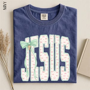 A navy blue t-shirt with a graphic design of the word "JESUS" in large letters, with a bow and ribbon decoration. The t-shirt is folded and placed on a surface, with some dried flowers and a book visible in the background.