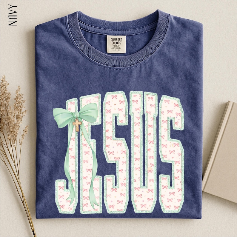 A navy blue t-shirt with a graphic design of the word "JESUS" in large letters, with a bow and ribbon decoration. The t-shirt is folded and placed on a surface, with some dried flowers and a book visible in the background.