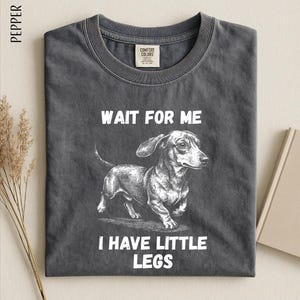 A gray t-shirt with a black and white illustration of a dachshund dog and the text "WAIT FOR ME I HAVE LITTLE LEGS" printed on it.