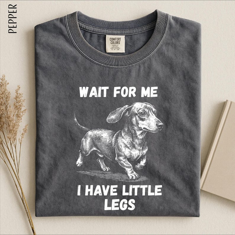 A gray t-shirt with a black and white illustration of a dachshund dog and the text "WAIT FOR ME I HAVE LITTLE LEGS" printed on it.
