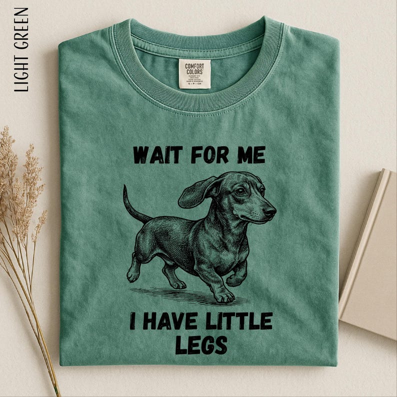 A green t-shirt with a black and white illustration of a dachshund dog and the text "WAIT FOR ME I HAVE LITTLE LEGS" printed on it.