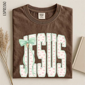 A brown t-shirt with the word "JESUS" printed on it, along with a bow and a small cross. The t-shirt is laid out on a surface, and there are some dried flowers and a book visible in the background.