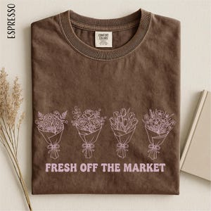 a brown t-shirt with a graphic design of four bouquets of flowers, each with a bow, and the text "FRESH OFF THE MARKET" printed on it.