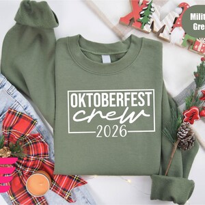 a green sweatshirt with the text "OKTOBERFEST CREW 2026" printed on it, along with various holiday-themed items such as a Christmas tree, ornaments, and a red bow.