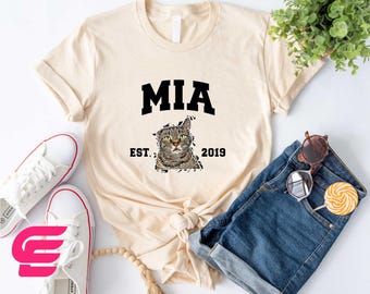 May include: A beige t-shirt with black text that reads "MIA EST. 2019". A cat's face is printed on the shirt.