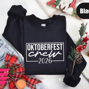a black sweatshirt with the text "OKTOBERFEST CREW" printed on it, along with a Christmas-themed background. The sweatshirt is placed on a white surface, surrounded by various holiday-related items such as a pair of jeans, a Christmas tree, and a candle.
