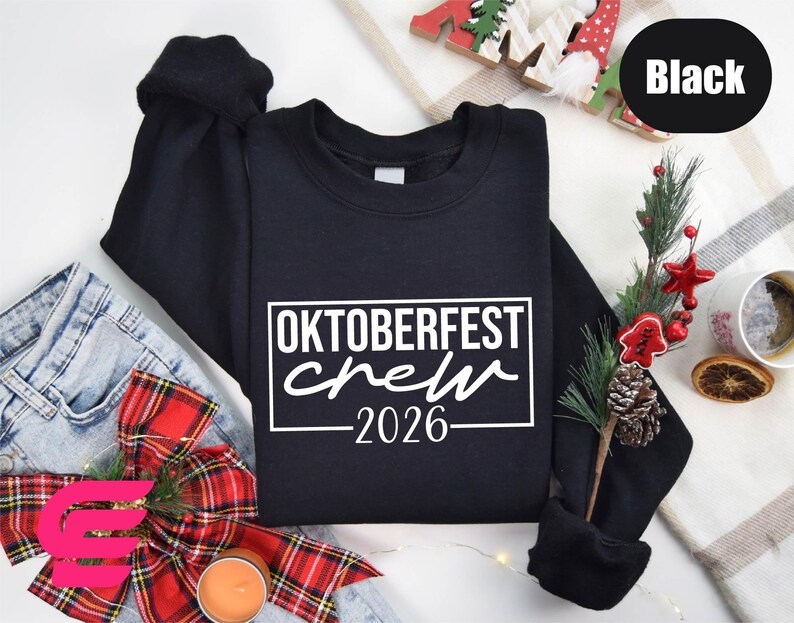 a black sweatshirt with the text "OKTOBERFEST CREW" printed on it, along with a Christmas-themed background. The sweatshirt is placed on a white surface, surrounded by various holiday-related items such as a pair of jeans, a Christmas tree, and a candle.